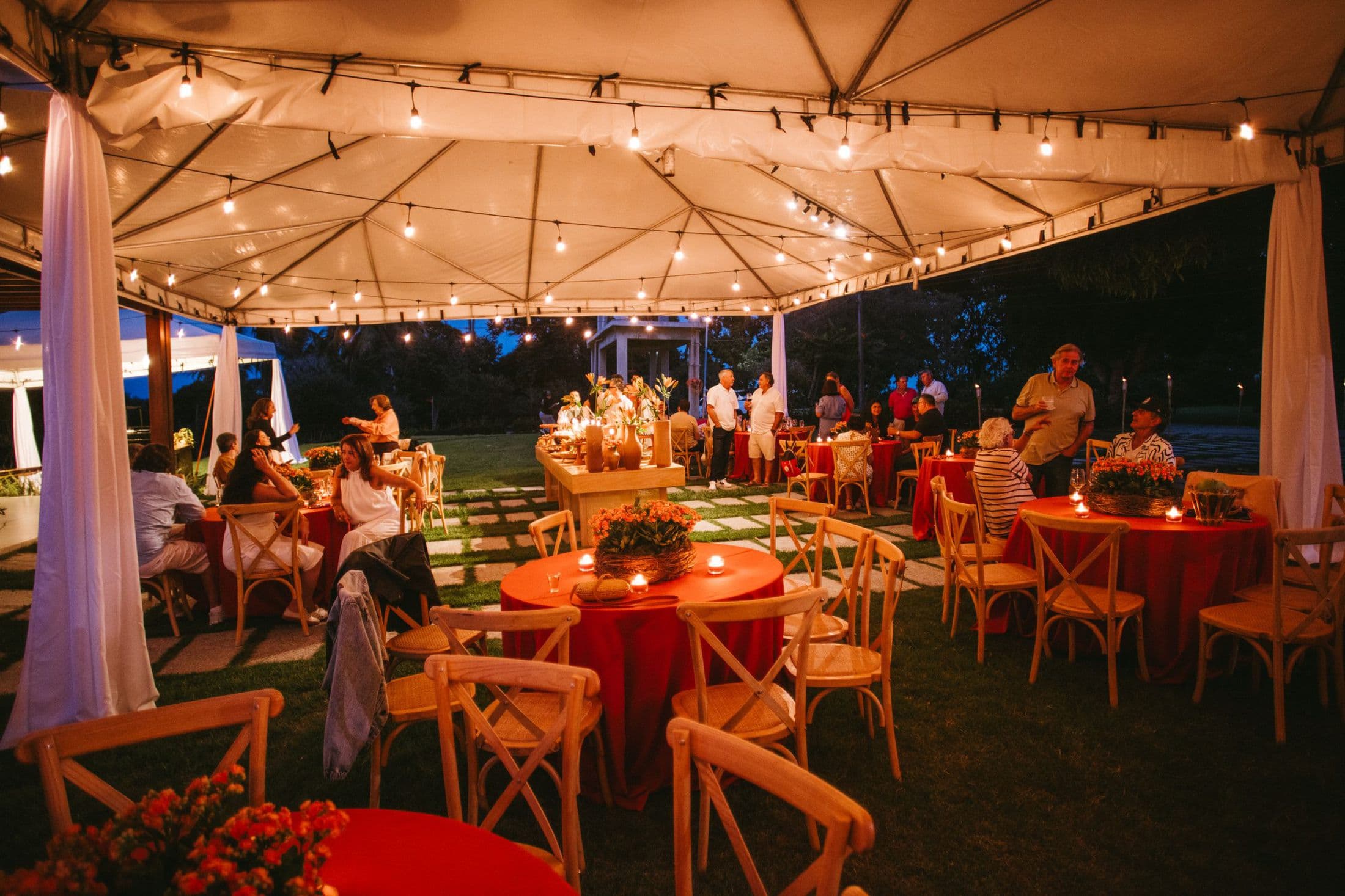Guests dining under a lit canopy tent in the evening with wooden crossback chairs